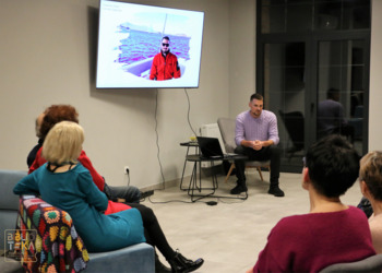 Mężczyzna siedzi przed grupą ludzi obok komputera i ekranu telewizora